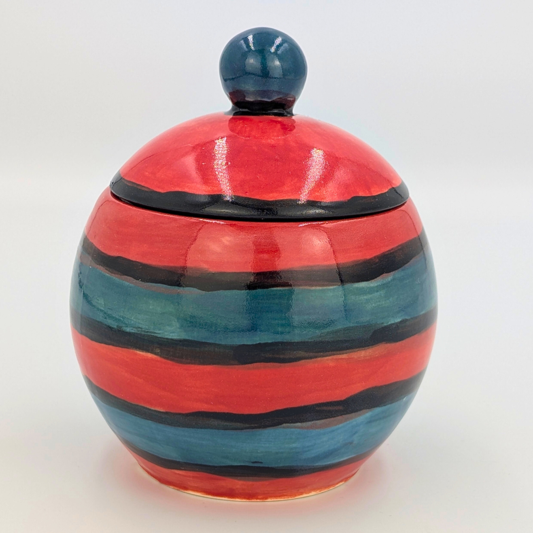 malowanie-ceramiki-gdansk-cukierniczka025l-paski-czerwono-niebieska Kolorowa cukierniczka o pojemności 0,25l z warsztatów malowania ceramiki w Gdańsku. Okrągły pojemnik ozdobiony poziomymi paskami w kolorach czerwonym, niebieskim i czarnym, z granatową gałką na pokrywce. Praktyczny i dekoracyjny element zastawy stołowej stworzony podczas warsztatów ceramicznych w Gdańsku.