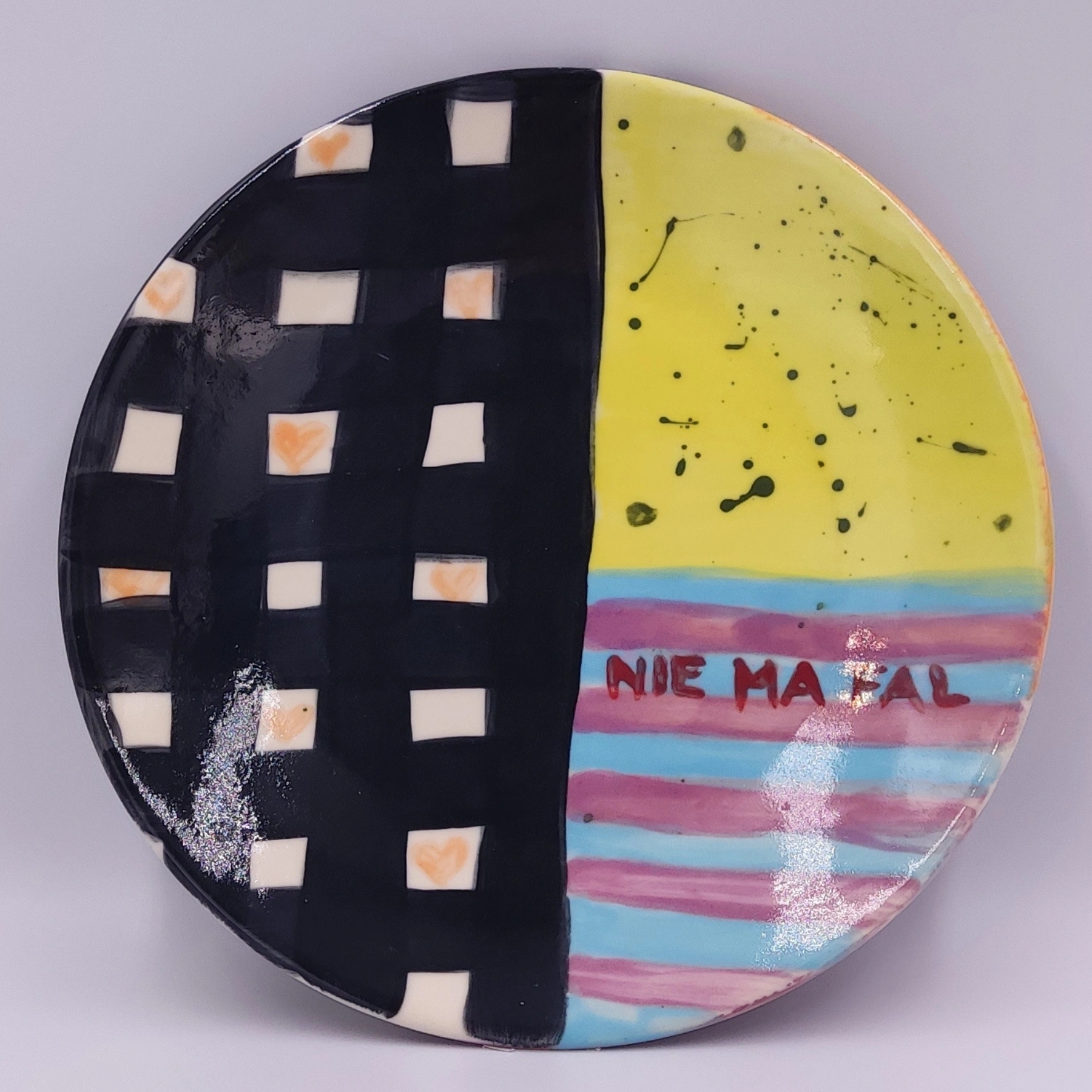 malowanie-ceramiki-gdansk-talerz19cm-kontrast-nie-ma-fal Artystyczny talerz ceramiczny z warsztatów malowania ceramiki w Gdańsku, podzielony na dwie części - jedną z czarno-białą kratką z pomarańczowymi akcentami, drugą kolorową z żółtym tłem, niebiesko-różowymi paskami i napisem "NIE MA FAL". Oryginalny projekt stworzony podczas warsztatów malowania ceramiki w Gdańsku.