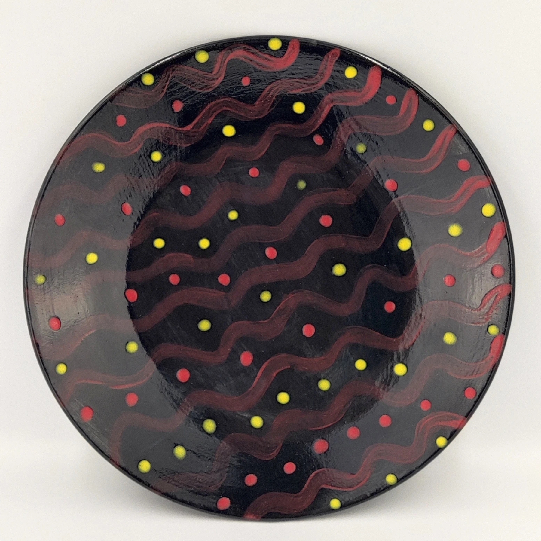 malowanie-ceramiki-gdansk-talerz25cm-czarny-fale-kropki Czarny talerz ceramiczny ozdobiony czerwonymi falistymi liniami i wzorem naprzemiennych czerwonych i żółtych kropek. Błyszcząca szkliwiona powierzchnia tworzy kontrast między ciemnym tłem a jaskrawymi elementami dekoracyjnymi.