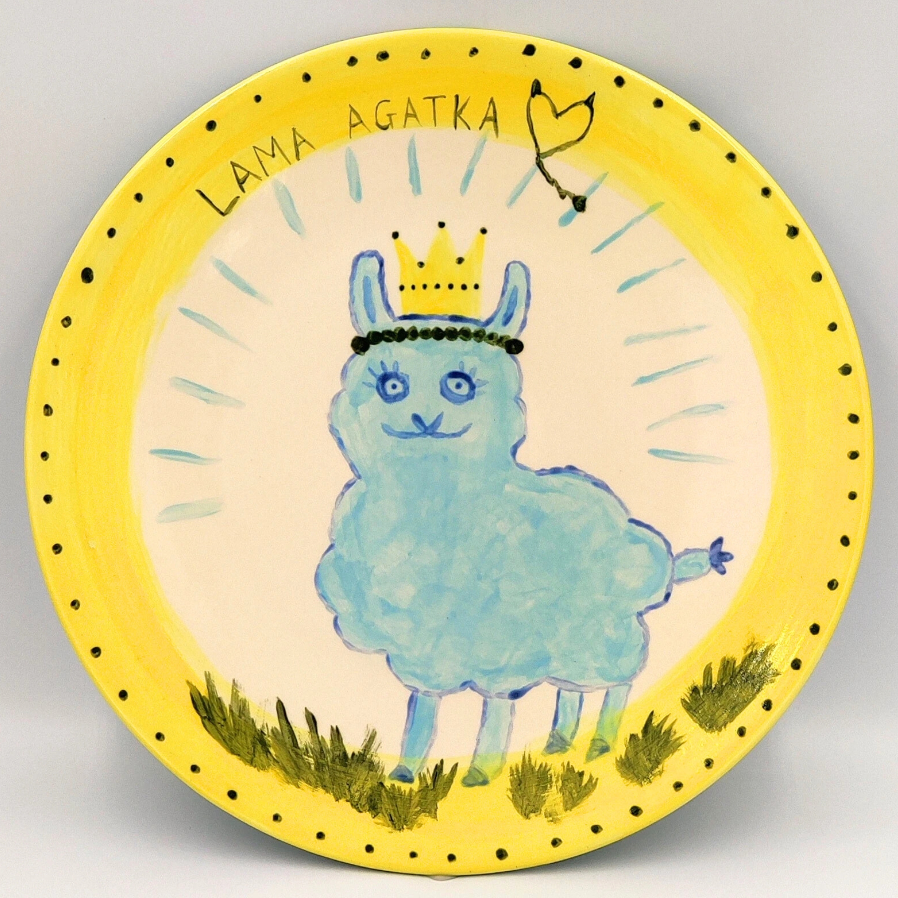 malowanie-ceramiki-gdansk-talerz25cm-lama-agatka-zolty Ręcznie malowany talerz ceramiczny z żółtym brzegiem i białym środkiem przedstawiający niebieską lamę w koronie. Talerz zawiera napis "LAMA AGATKA" i serduszko na górze, czarne kropki wokół krawędzi oraz zieloną trawę na dole.